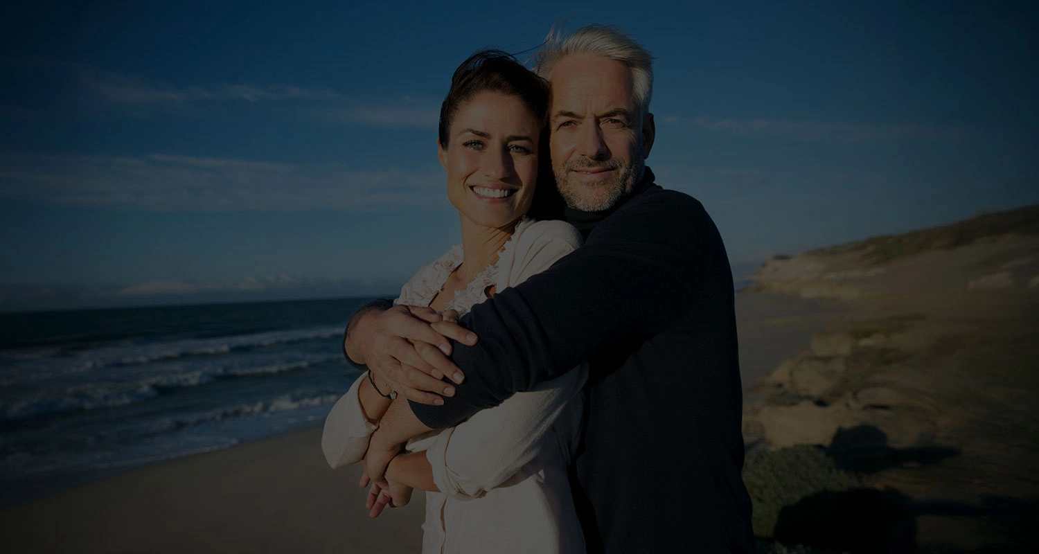 Happy retired couple cuddling along the beach at sunset.